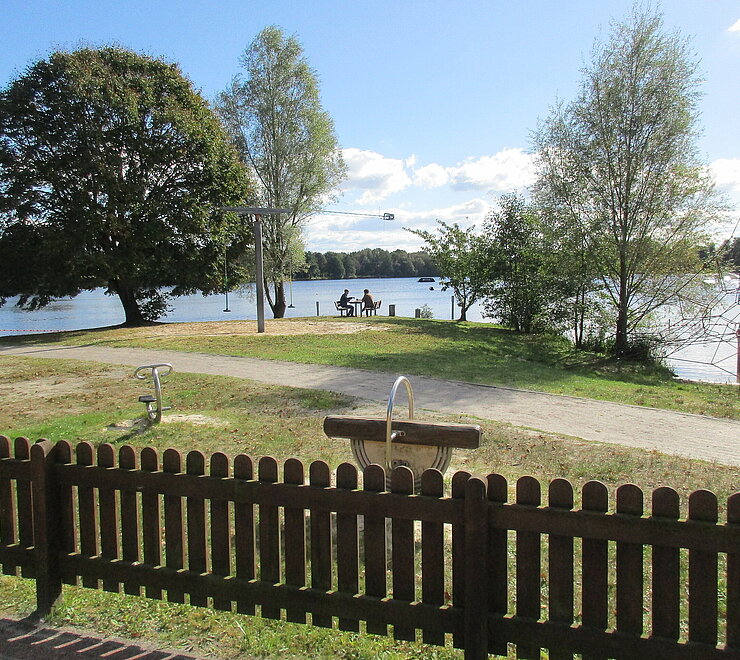 Blick auf den Idasee. Vorne steht ein kleiner brauner Gartenzaun. Im Hintergrund stehen Spielgeräte und eine Bank wo zwei Personen sitzen. 