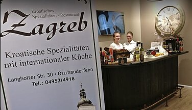 Das Bild zeigt den Eingangsbereich des Restaurant Zagreb. Zwei Damen stehen an Empfang. Im Vordergrund steht ein großes Schild auf dem die Adresse des Restaurants geschrieben ist ebenso welche Richtung der Speisen angeboten wird.