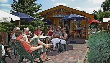 Das Bild zeigt das Café Melkuske an einem Sommertag. Mehrere Personen sitzen vor dem in Holzoptik erlassenen Café und trinken ein Getränk. Mehrere Sonnenschirme stehen zum Schutz um die Tische herum. Im Hintergrund sieht man die Hütte des Café's und den Eingang.