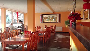 Auf dem Bild ist der Innenraum des Restaurant Asia Wok. Rechts an der Seite steht die Theke des Restaurants, in der Mitte des Raumes stehen mehrere Tischpartien unterschiedlicher Stuhlanzahl. Geradeaus an der Wand hängt ein Bild im Chinesischen style. Das Restaurant ist Dekoriert mit typischen Chinesischen Dekoartikeln und kulturellen Gegenständen..