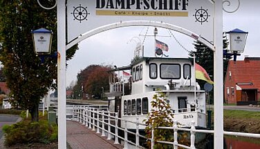 Auf dem Bild ist das Eingangstor der Kneipe ,,Dampfschiff''. Im Hintergrund liegt das weiße Schiff mit Fahnen die das äußere des Schiffs farblich schmücken.