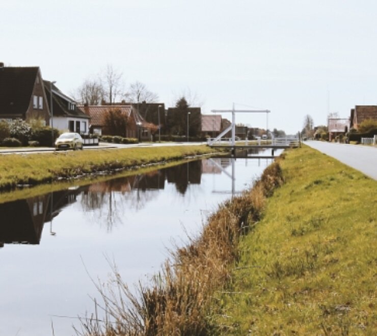 Das Foto entstand in der 1.Süwieke. In der Mitte des Bildes fließt der Kanal entlang. Im Hintergrund steht eine weiße Fehnbrücke und an den Seiten des Kanals wächst Schilf. 