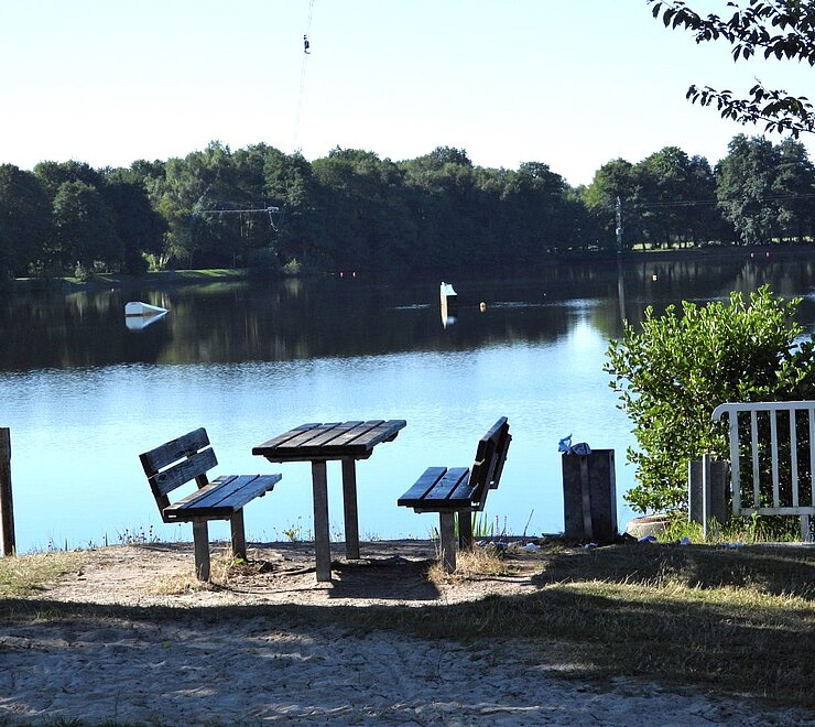 Eine Picknick-Bank steht vor dem Idasee. Im Hintergrund sieht man den See und ihre  Wasserskianlage. 