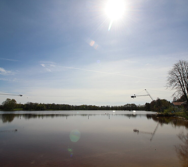 Blick auf den Idasee bei strahlendem Sonnenschein, in Mitten des Idasees stehen metallische Gerüste um die Wasserskianlage zu betreiben.